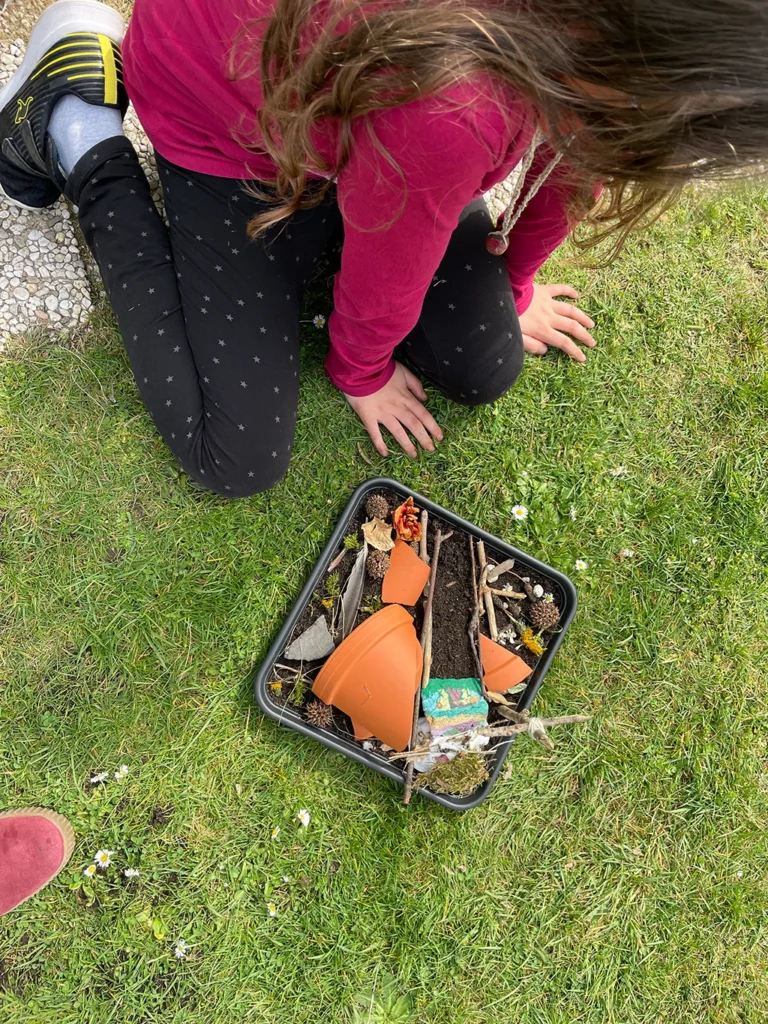 Kind mit einer Schale, in der ein Minigarten mit Erde, kleinen Ästen und Tonscherben ist.