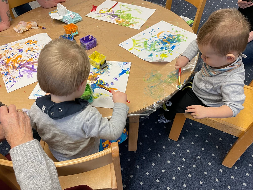 Die Kleinkinder sitzen am Tische und malen mit vielen bunten Farben.